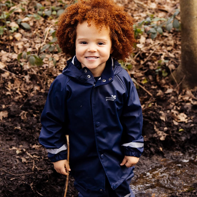 A young child smiling and wearing a Rainy Day Waterproof Jacket Navy in a woodland setting.